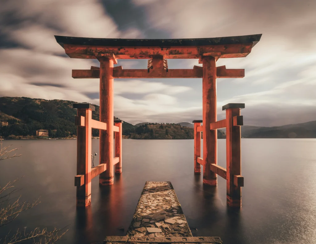 Vermilion torii gate standing in lake water at a stone path, distant mountains and soft clouds filling the horizon.