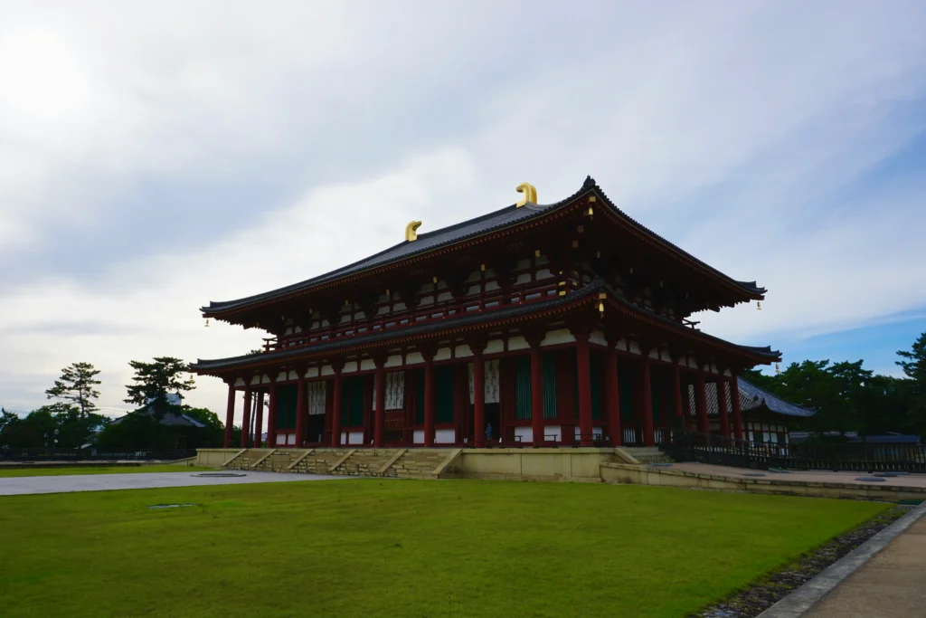 Large wooden temple hall with red columns, tiered tiled roof and gold details beside an open lawn under bright sky.