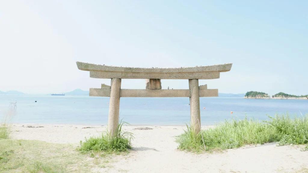 Stone torii gate on a sandy beach facing a calm blue bay, with distant islets and a ship on the horizon.