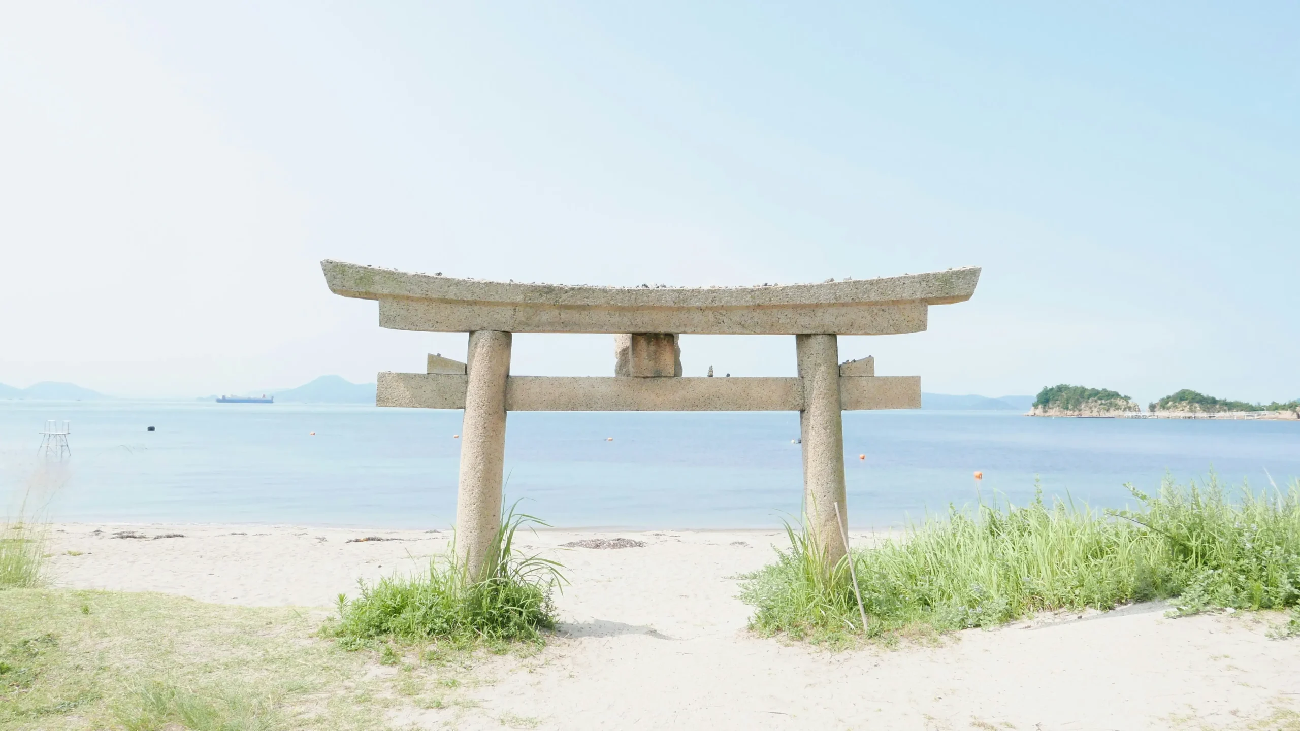 Stone torii gate on a sandy beach facing a calm blue bay, with distant islets and a ship on the horizon.