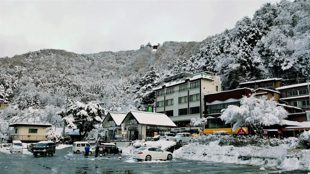 Snow-covered Japanese town below forested hills, cars and ryokan around a small station square after fresh snowfall.