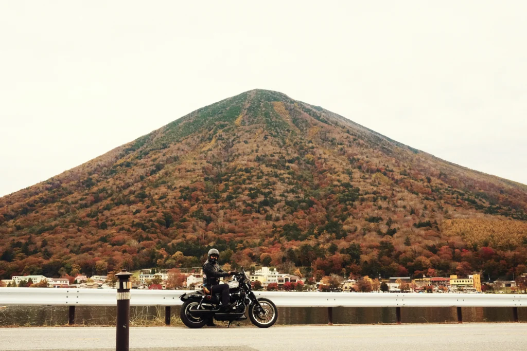 Black cruiser motorcycle at guardrail; conical forested mountain and lakeside town behind, autumn colors under overcast sky