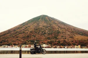 Black cruiser motorcycle at guardrail; conical forested mountain and lakeside town behind, autumn colors under overcast sky