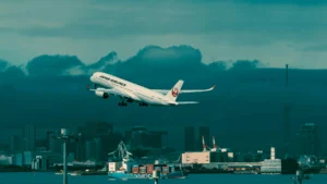 Japan Airlines jet climbing over Tokyo Bay with skyline and port cranes—shot typical of Haneda departures.