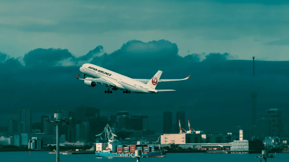 Japan Airlines jet climbing over Tokyo Bay with skyline and port cranes—shot typical of Haneda departures.