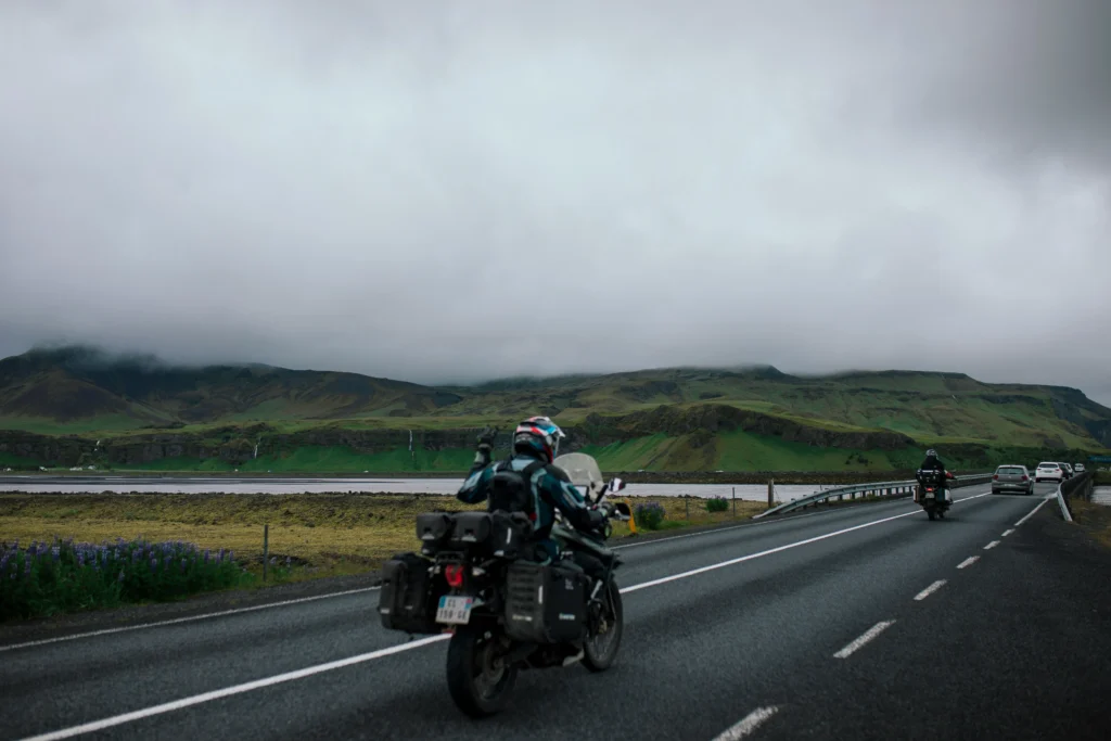 Two touring motorcyclists ride a lakeside highway toward misty green hills under low clouds, with cars and roadside flowers.