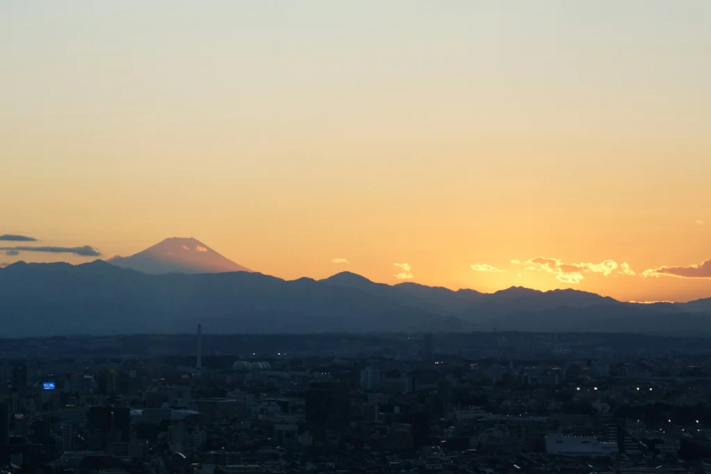 Distant silhouette of Mount Fuji behind layered hills and a wide city skyline under an orange dusk horizon.
