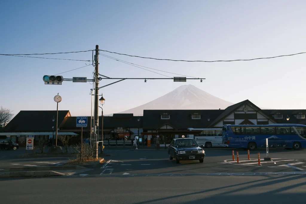 Mount Fuji behind Kawaguchiko Station at dusk, with wires, green light, tour buses and a black taxi in the forecourt.