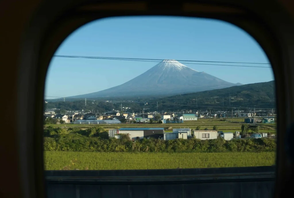 Train window frames distant Mount Fuji beyond green fields and small town buildings, with thin power lines crossing the view.