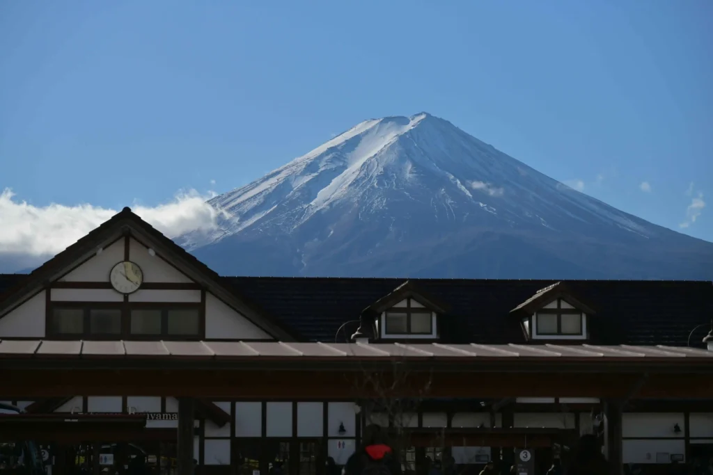 Mount Fuji under a clear blue sky above Kawaguchiko Station’s gabled roof, with a round clock centered on the facade.