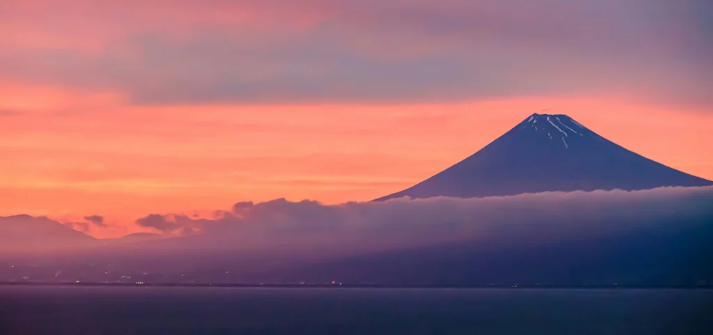 Mount Fuji at dawn with a pink sky, snow streaks on the summit, a low cloud band across the slope, and still water in the foreground.