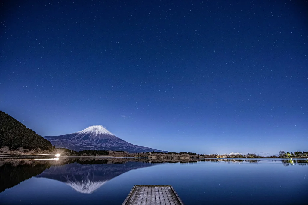 A glassy lake mirrors Mount Fuji under a field of stars. Peaceful, clear-sky conditions ideal for long-exposure shots from a quiet pier along the Fuji Five Lakes.