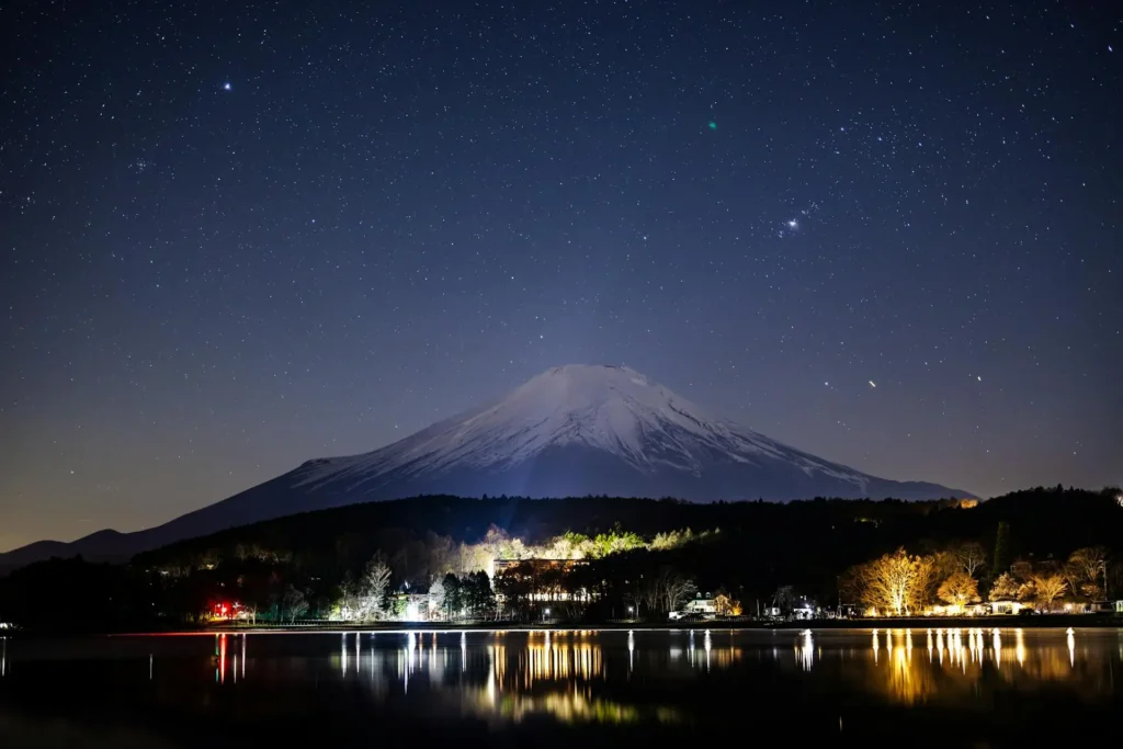 Mount Fuji rises over a lakeside town glowing with warm lights, its snowy slopes set against a sparkling sky—an atmospheric scene for winter travel dreams.