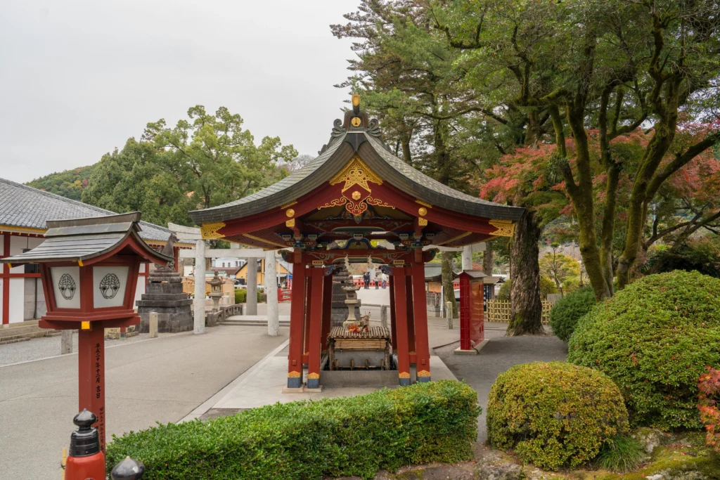 Red and gold shrine pavilion with water basin between stone torii and lanterns, bordered by maples in a calm temple courtyard.