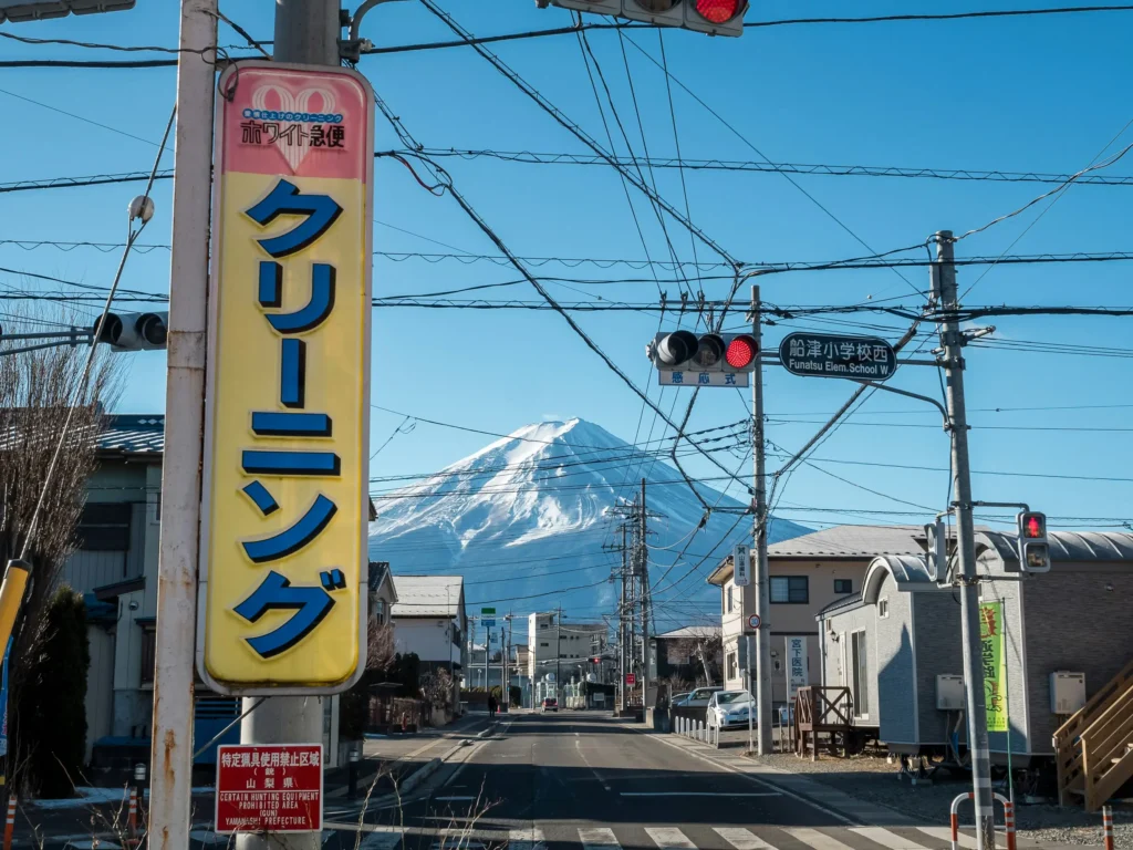 Small-town intersection with Japanese signboards, traffic lights, and overhead wires; Mt. Fuji centered in the distance