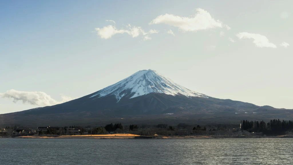 Admire Mount Fuji’s snow-capped peak under clear skies from Lake Yamanaka — a peaceful setting where light clouds and golden shores highlight Japan’s natural splendor.