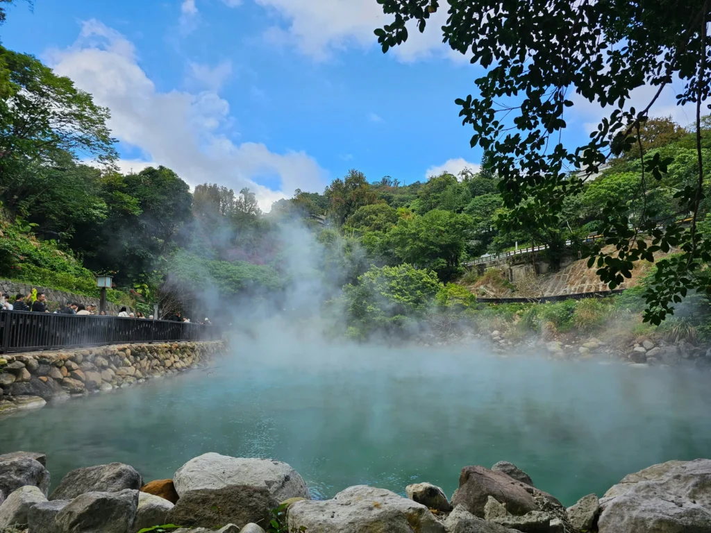 A steaming natural hot spring with visitors observing from a walkway, framed by trees and a clear blue sky.